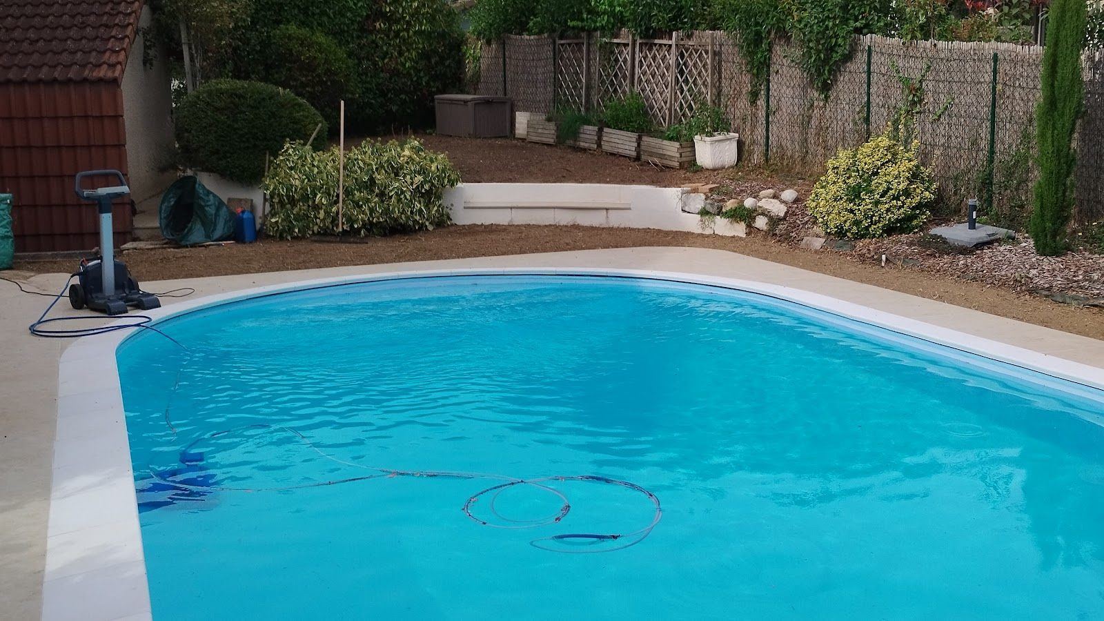 Abords d'une piscine : terre préparée entre le bassin et la clôture, arbustes et cyprès conservés